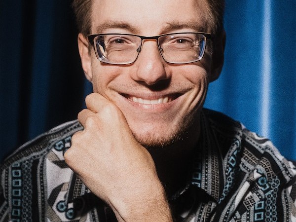 Ein Mann mit Brille lächelt in die Kamera. Er trägt ein gemustertes Hemd und sitzt vor einem blauen Vorhang., © Kulturhaus Schwanen