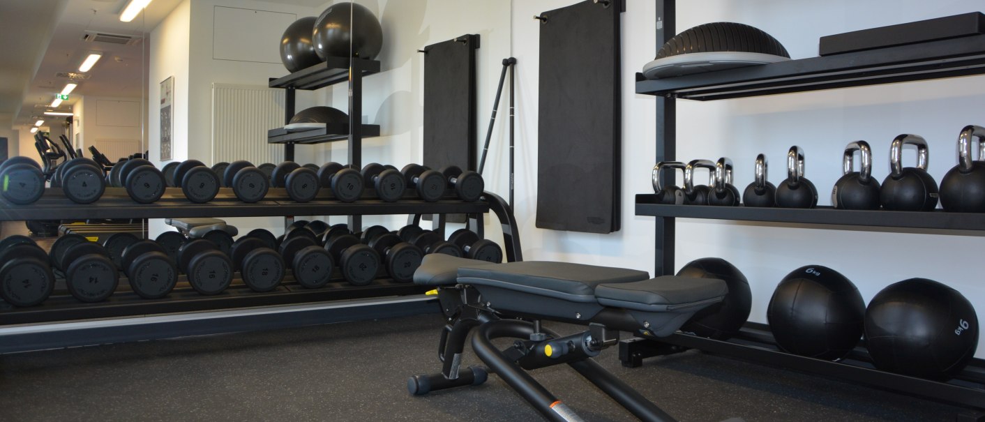 Ein modernes Fitnessstudio mit Hanteln, Kettlebells und Gymnastikb&auml;llen. Eine Trainingsbank steht im Vordergrund, Spiegel an der Wand., &copy; Aloft Stuttgart