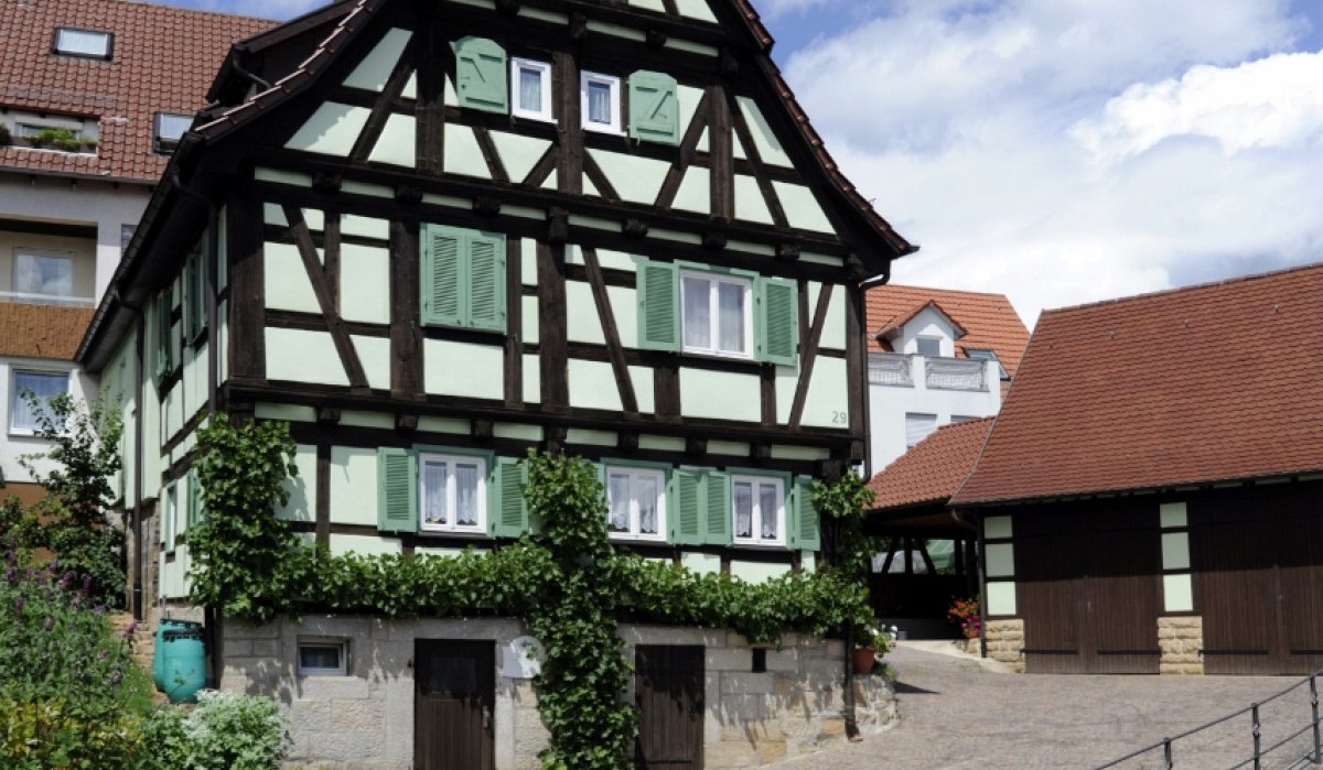Fachwerkhaus mit grünem Anstrich und blauen Fensterläden, umgeben von einem gepflasterten Hof und blühenden Pflanzen, unter blauem Himmel., © Natur.Nah. Schönbuch & Heckengäu Fachwerkhaus mit grünem Anstrich und blauen Fensterläden, umgeben von einem gepflasterten Hof und blühenden Pflanzen, unter blauem Himmel., © Natur.Nah. Schönbuch & Heckengäu