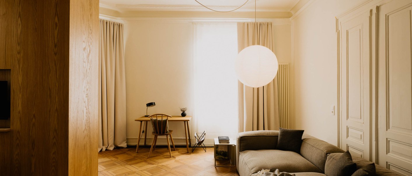 Elegantes Wohnzimmer mit einem grauen Sofa, Holzboden, Schreibtisch und großer runder Hängelampe. Helle Vorhänge und minimalistische Einrichtung., © Peter Oliver Wolff / DAS EDITH