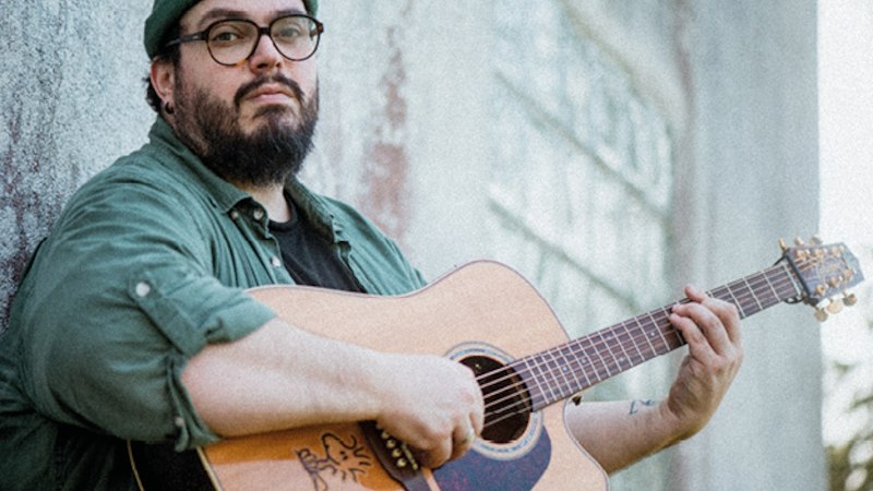 Ein Mann mit Brille und Mütze spielt Gitarre und lehnt an einer Wand. Er trägt ein grünes Hemd und schaut in die Kamera., © IMK - Rainer Koczwara Ein Mann mit Brille und Mütze spielt Gitarre und lehnt an einer Wand. Er trägt ein grünes Hemd und schaut in die Kamera., © IMK - Rainer Koczwara