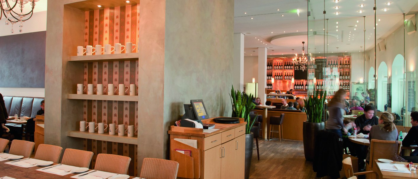 Stilvolles Restaurant mit Holzmöbeln, Regalen mit Tassen und einer Bar im Hintergrund. Menschen sitzen an Tischen, moderne Beleuchtung und Pflanzen dekorieren den Raum., © aspectus photographie, Stuttgart