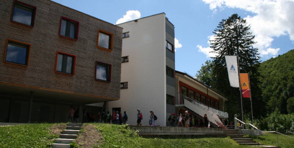 Moderne Jugendherberge in Bad Urach mit Holzfassade und bunten Fenstern, umgeben von grüner Natur und blauem Himmel.