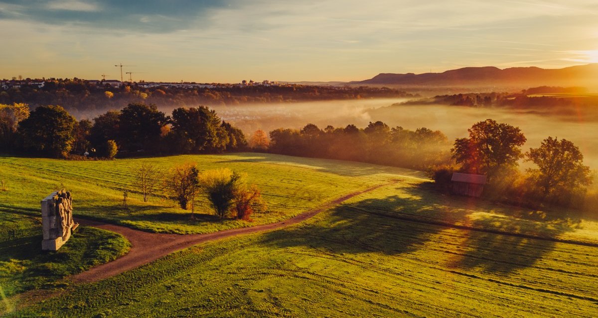 Sonnenaufgang über grünen Feldern mit Bäumen, einem Kunstwerk und Nebel in der Ferne. Die Sonne wirft lange Schatten., © Landratsamt Ostalbkreis Sonnenaufgang über grünen Feldern mit Bäumen, einem Kunstwerk und Nebel in der Ferne. Die Sonne wirft lange Schatten., © Landratsamt Ostalbkreis