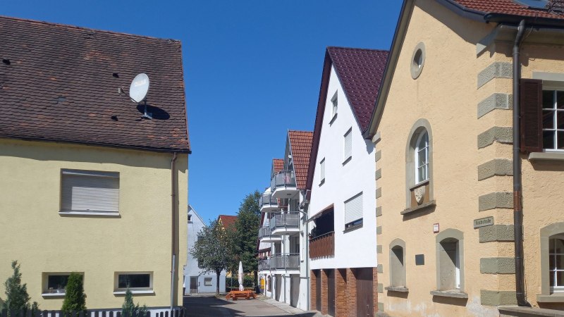 Eine ruhige Straße mit Häusern und einem klaren blauen Himmel. Die Gebäude haben rote Ziegeldächer und helle Fassaden., © Petra Natzkowski Eine ruhige Straße mit Häusern und einem klaren blauen Himmel. Die Gebäude haben rote Ziegeldächer und helle Fassaden., © Petra Natzkowski