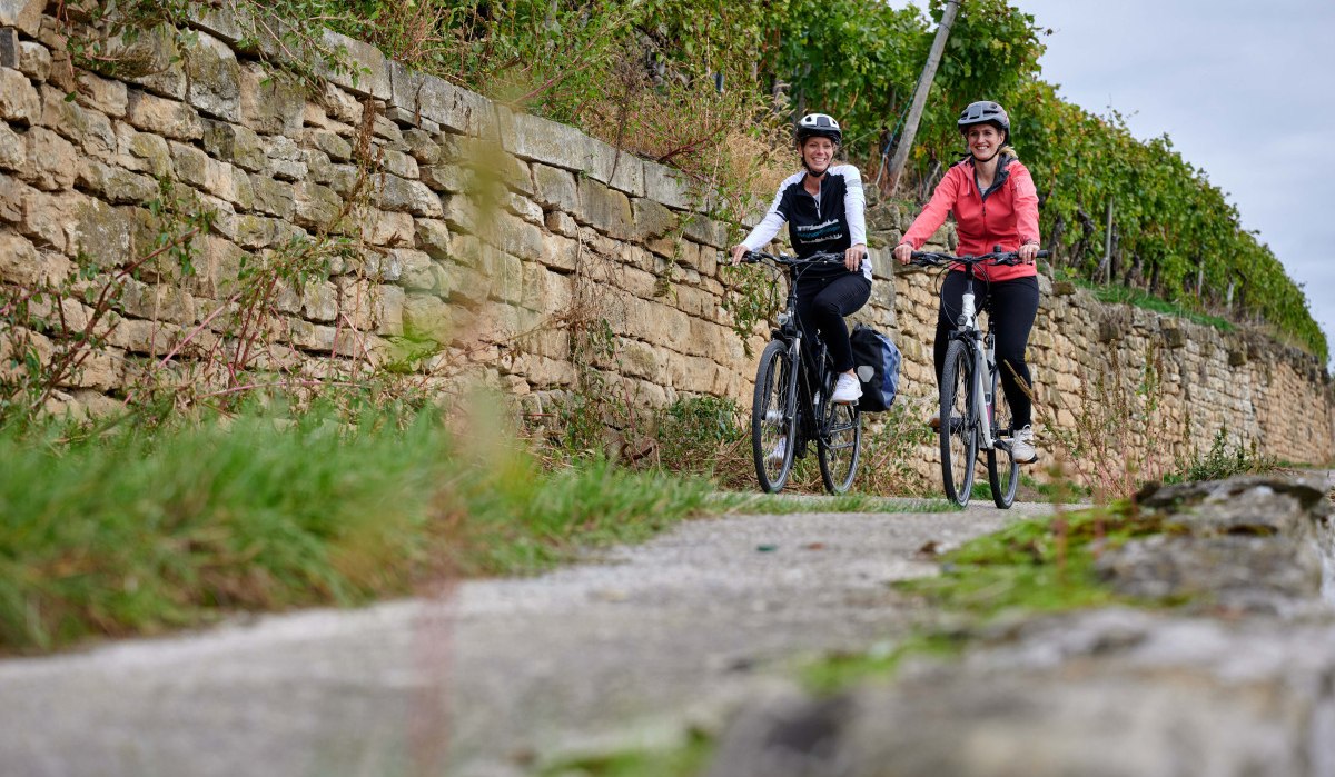 Zwei Frauen radeln lächelnd auf einem Weg neben einer alten Steinmauer, umgeben von grüner Vegetation., © Kraichgau Stromberg Tourismus e.V.-Christian Ernst Zwei Frauen radeln lächelnd auf einem Weg neben einer alten Steinmauer, umgeben von grüner Vegetation., © Kraichgau Stromberg Tourismus e.V.-Christian Ernst