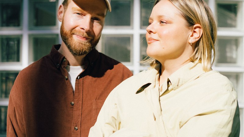 Zwei Personen vor einer Glaswand, eine trägt eine Mütze und ein braunes Hemd, die andere eine helle Jacke. Beide lächeln in die Sonne., © Scala Kultur Live gGmbH Zwei Personen vor einer Glaswand, eine trägt eine Mütze und ein braunes Hemd, die andere eine helle Jacke. Beide lächeln in die Sonne., © Scala Kultur Live gGmbH