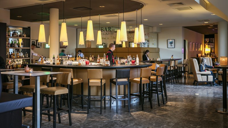 Moderne Bar mit runder Theke, Hängelampen und Sitzbereichen im Arcotel Camino. Menschen sitzen und genießen die Atmosphäre., © Harald Eisenberger Moderne Bar mit runder Theke, Hängelampen und Sitzbereichen im Arcotel Camino. Menschen sitzen und genießen die Atmosphäre., © Harald Eisenberger