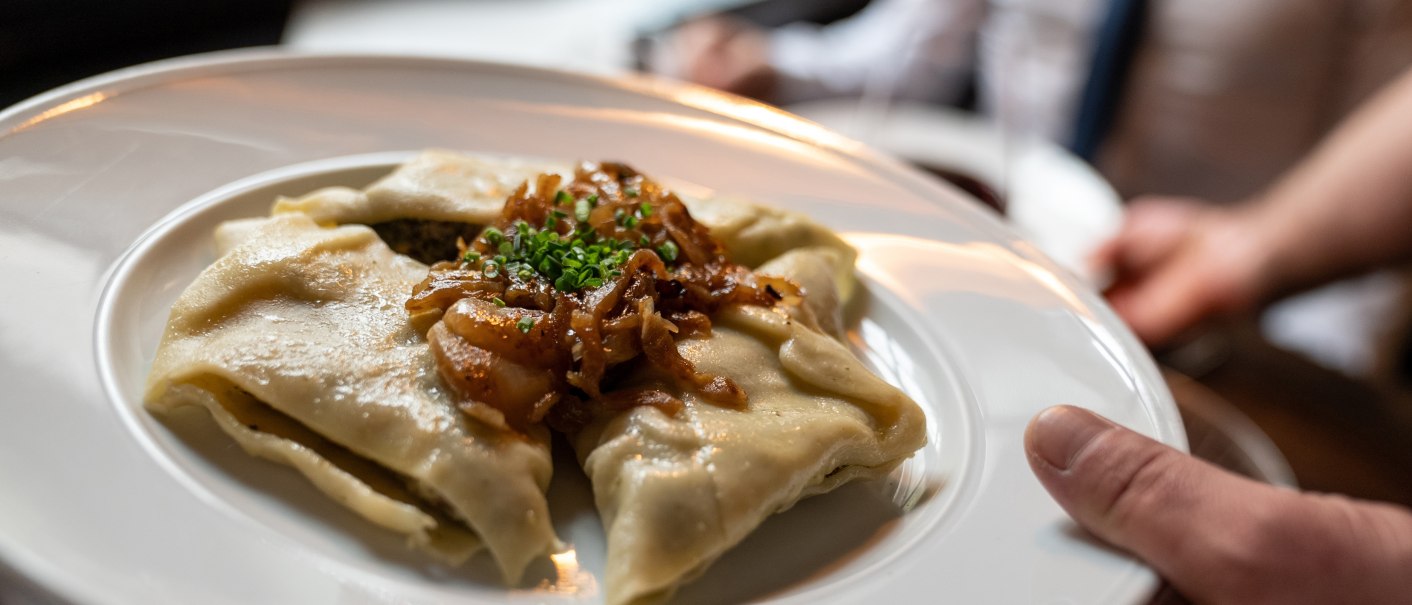 Ein Teller mit Maultaschen, garniert mit geschmolzenen Zwiebeln und Schnittlauch, wird serviert. Im Hintergrund ist eine Person unscharf zu sehen., © Stuttgart-Marketing GmbH, Martina Denker