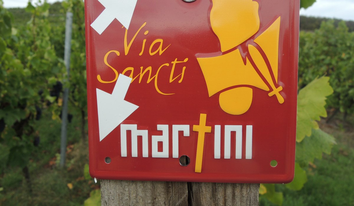 Rotes Schild mit 'Via Sancti Martini' und Pfeilen, befestigt an einem Holzpfosten in einem Weinberg., © Land der 1000 Hügel - Kraichgau-Stromberg Rotes Schild mit 'Via Sancti Martini' und Pfeilen, befestigt an einem Holzpfosten in einem Weinberg., © Land der 1000 Hügel - Kraichgau-Stromberg