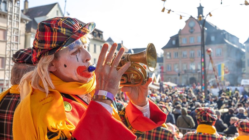 Ein Clown mit bemaltem Gesicht und bunter Kleidung spielt Trompete auf einem belebten Karnevalsumzug vor einem historischen Geb&auml;ude., &copy; F&uuml;r die Bildrechte: Amt f&uuml;r Medien und Kommunikation Schw&auml;bisch Gm&uuml;nd