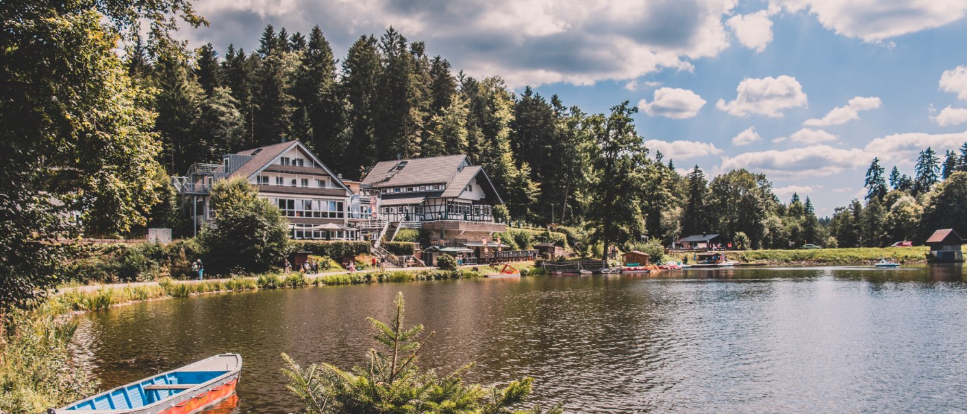 Ein idyllischer See mit einem Boot im Vordergrund, umgeben von Wald. Am Ufer steht ein großes Gebäude unter blauem Himmel mit Wolken., © Stadt Welzheim