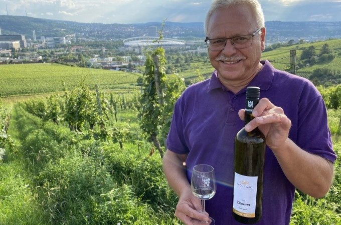 Ein Mann in einem lila Hemd hält eine Weinflasche und ein Glas in einem Weinberg. Im Hintergrund ist Stuttgart mit einem Stadion zu sehen., © Weingenuss Stuttgart