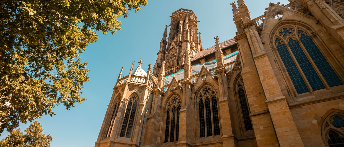 Die Johanneskirche in Stuttgart mit gotischer Architektur, umgeben von Bäumen und strahlend blauem Himmel., © SMG, Sarah Schmid Die Johanneskirche in Stuttgart mit gotischer Architektur, umgeben von Bäumen und strahlend blauem Himmel., © SMG, Sarah Schmid