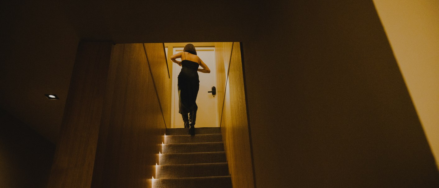Eine Person in einem dunklen Kleid geht eine beleuchtete Treppe hinauf zu einer Tür in einem schmalen Flur., © Peter Oliver Wolff / DAS EDITH