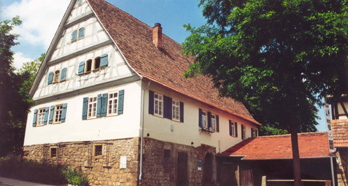 Fachwerkhaus mit rotem Dach und blauen Fensterläden, umgeben von Bäumen. Das Gebäude steht auf einem steinernen Fundament., © Alfred Rösner