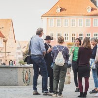 Eine Gruppe von Menschen steht um einen Marktbrunnen in einer Altstadt mit Fachwerkhäusern. Ein Mann in Hut und Mantel scheint eine Führung zu geben., © Bebop Media, Danijel Grbic