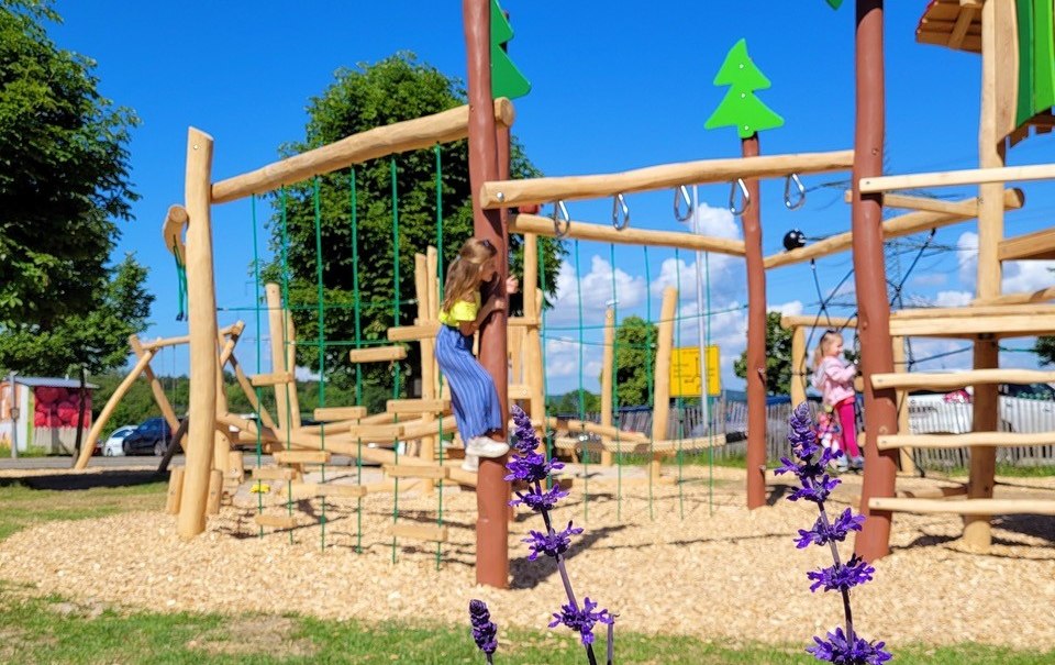 Ein Spielplatz mit Klettergerüst aus Holz, umgeben von bunten Blumen. Kinder spielen im Hintergrund. Der Himmel ist klar und blau., © Engel/ Rammerthof