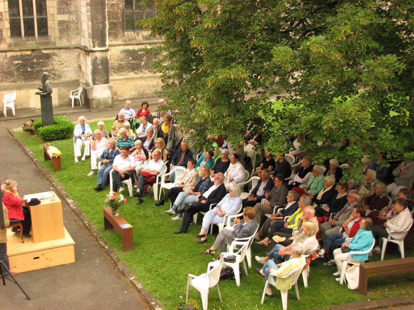 Menschen sitzen im Freien auf wei&szlig;en St&uuml;hlen und h&ouml;ren einer Lesung zu. Die Veranstaltung findet in einem historischen Hof statt, umgeben von B&auml;umen., &copy; Kulturreferat Bad Urach