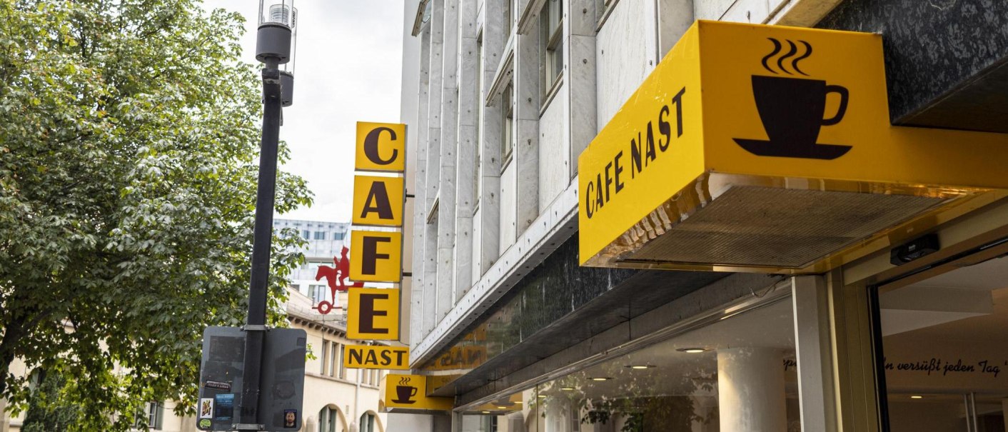 Gelbes Schild des Cafe Nast mit Kaffeetasse-Logo an einem Gebäude, umgeben von Bäumen., © SMG, Sarah Schmid Gelbes Schild des Cafe Nast mit Kaffeetasse-Logo an einem Gebäude, umgeben von Bäumen., © SMG, Sarah Schmid