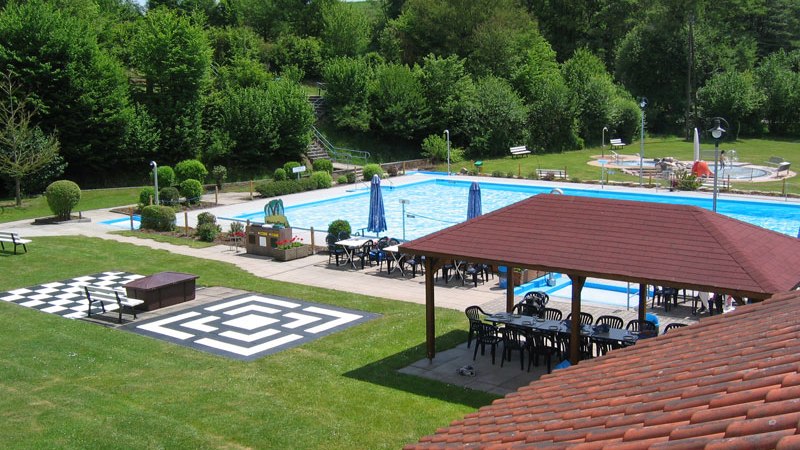 Ein Freibad mit Schwimmbecken, umgeben von Bäumen. Es gibt Sitzbereiche mit Sonnenschirmen und einen überdachten Pavillon., © FVFS Ein Freibad mit Schwimmbecken, umgeben von Bäumen. Es gibt Sitzbereiche mit Sonnenschirmen und einen überdachten Pavillon., © FVFS