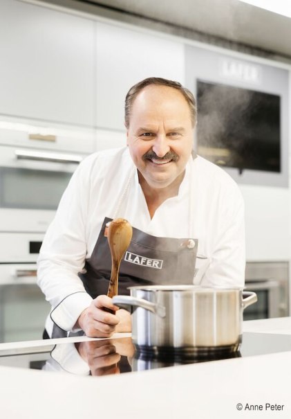 Ein Koch in einer modernen K&uuml;che r&uuml;hrt mit einem Holzl&ouml;ffel in einem Topf. Er tr&auml;gt eine Sch&uuml;rze mit der Aufschrift 'Lafer'., &copy; Stadthalle Leonberg