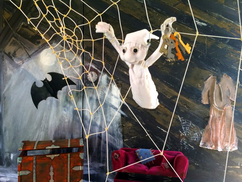 Ein kleines Gespenst hängt in einem Spinnennetz, hält Schlüssel. Im Hintergrund eine Fledermaus, eine offene Truhe und ein rotes Sofa., © Stadtbücherei Bad Urach