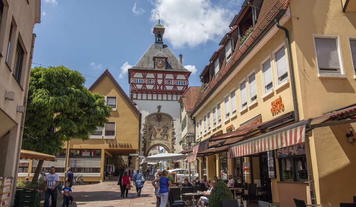 Straßenszene in einer Altstadt mit Fachwerkhaus und Uhrturm. Menschen flanieren und sitzen in Straßencafés. Der Himmel ist blau mit wenigen Wolken., © Stuttgart Marketing GmbH Achim Mende