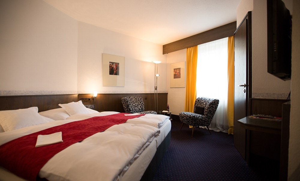 Gem&uuml;tliches Hotelzimmer mit Doppelbett, roten Akzenten, zwei Sesseln, gelben Vorh&auml;ngen und einem Wandfernseher., &copy; Hotel B&uuml;rkle
