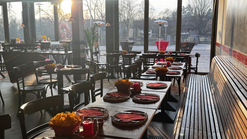 Ein Restaurant mit gedeckten Tischen, roten Servietten und Kerzen. Sonnenlicht strömt durch große Fenster und beleuchtet den Raum., © JoePenas Ein Restaurant mit gedeckten Tischen, roten Servietten und Kerzen. Sonnenlicht strömt durch große Fenster und beleuchtet den Raum., © JoePenas