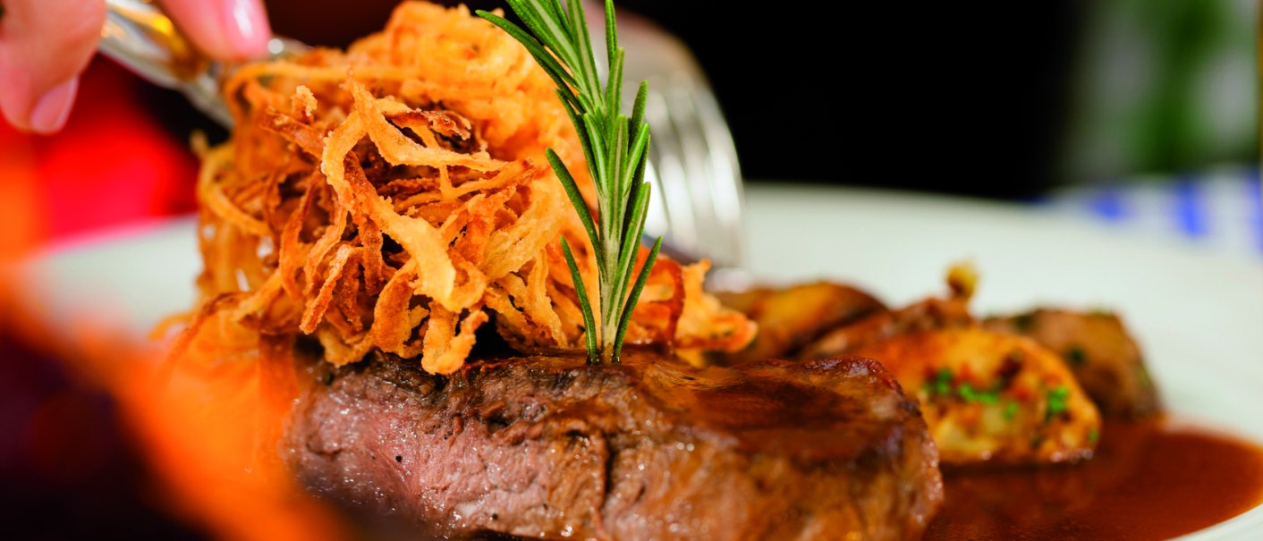 Zwiebelrostbraten mit knusprigen Röstzwiebeln und Rosmarin auf einem Teller, im Hintergrund unscharf gedeckter Tisch., © Fotolia, Kzenon Zwiebelrostbraten mit knusprigen Röstzwiebeln und Rosmarin auf einem Teller, im Hintergrund unscharf gedeckter Tisch., © Fotolia, Kzenon