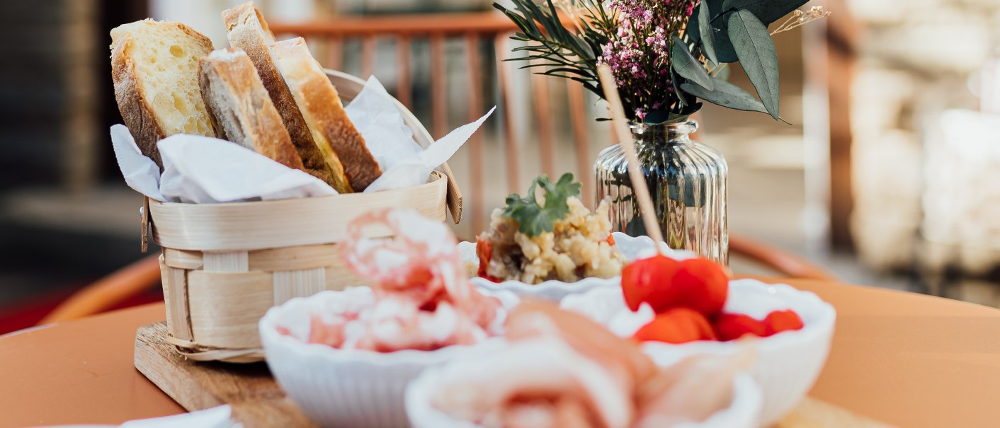 Ein Tisch im Freien mit Antipasti, Brot und Blumen in einem Café. Im Hintergrund sind Fenster und eine geöffnete Tür zu sehen., © L. Prestel/ Documenting Love Ein Tisch im Freien mit Antipasti, Brot und Blumen in einem Café. Im Hintergrund sind Fenster und eine geöffnete Tür zu sehen., © L. Prestel/ Documenting Love