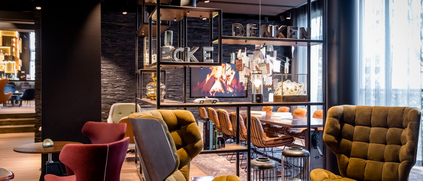 Moderne Lounge mit gemütlichen Sesseln, Regalen und einem Bildschirm mit Kaminfeuer. Schriftzug "DENKEN" auf dem Regal., © TOMAS