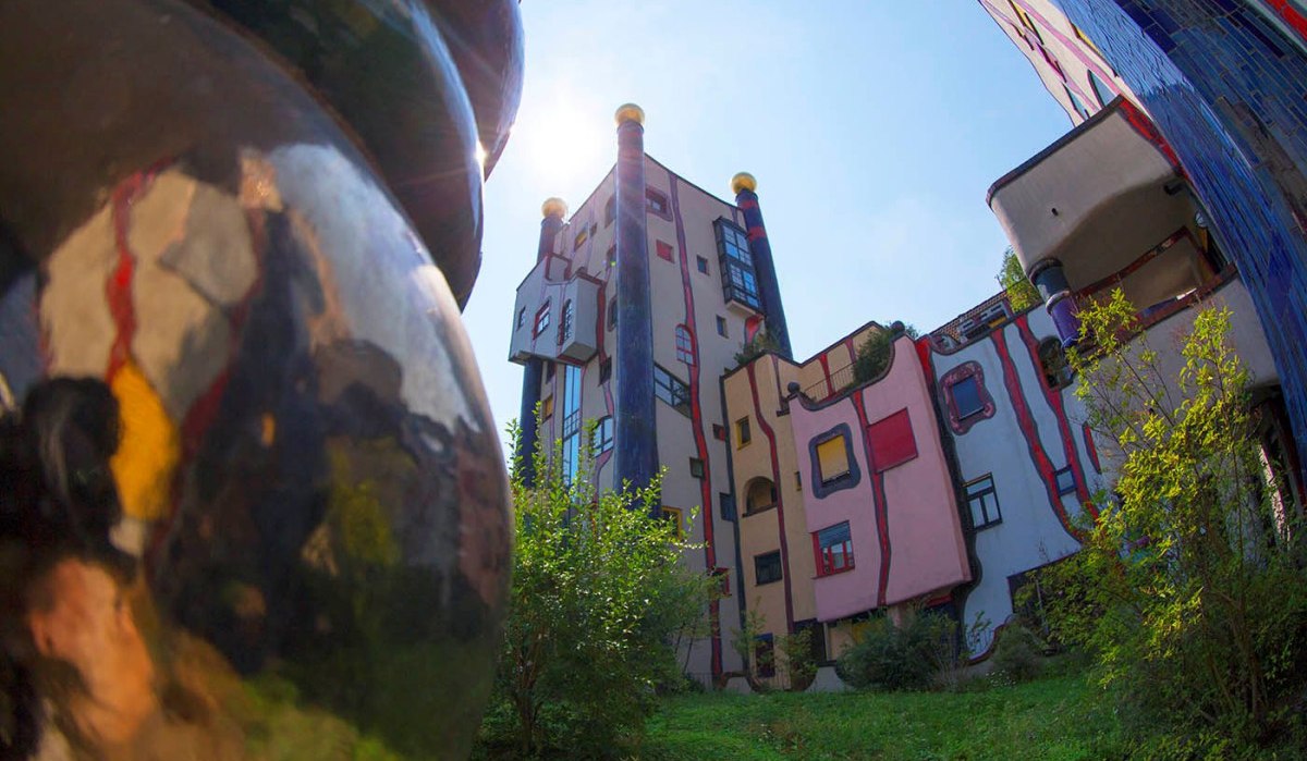 Bunte, unregelmäßige Gebäude mit runden Formen und goldenen Kuppeln im Hundertwasser-Stil, umgeben von Grünflächen., © Plochingen