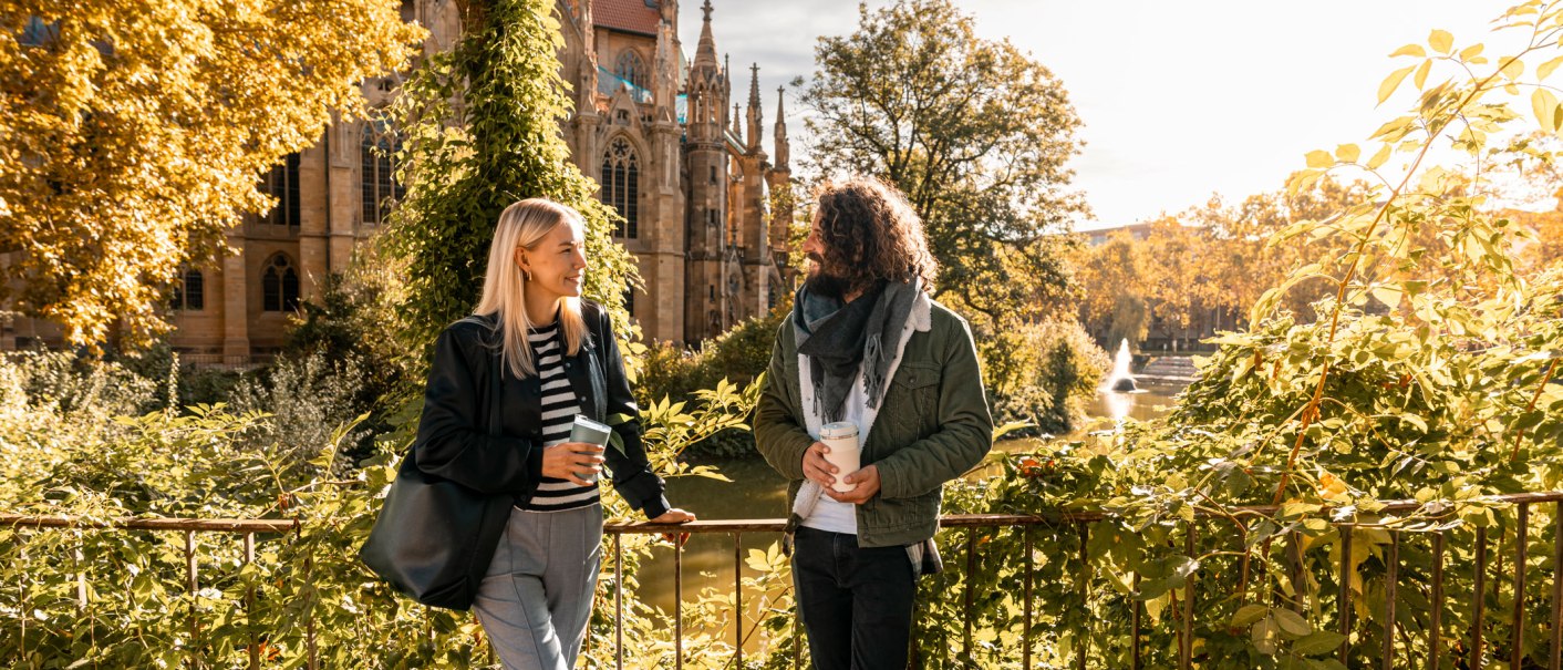 Zwei Personen unterhalten sich vor der Johanneskirche am Feuersee, umgeben von herbstlichem Laub und Sonnenschein., © SMG, Sarah Schmid Zwei Personen unterhalten sich vor der Johanneskirche am Feuersee, umgeben von herbstlichem Laub und Sonnenschein., © SMG, Sarah Schmid