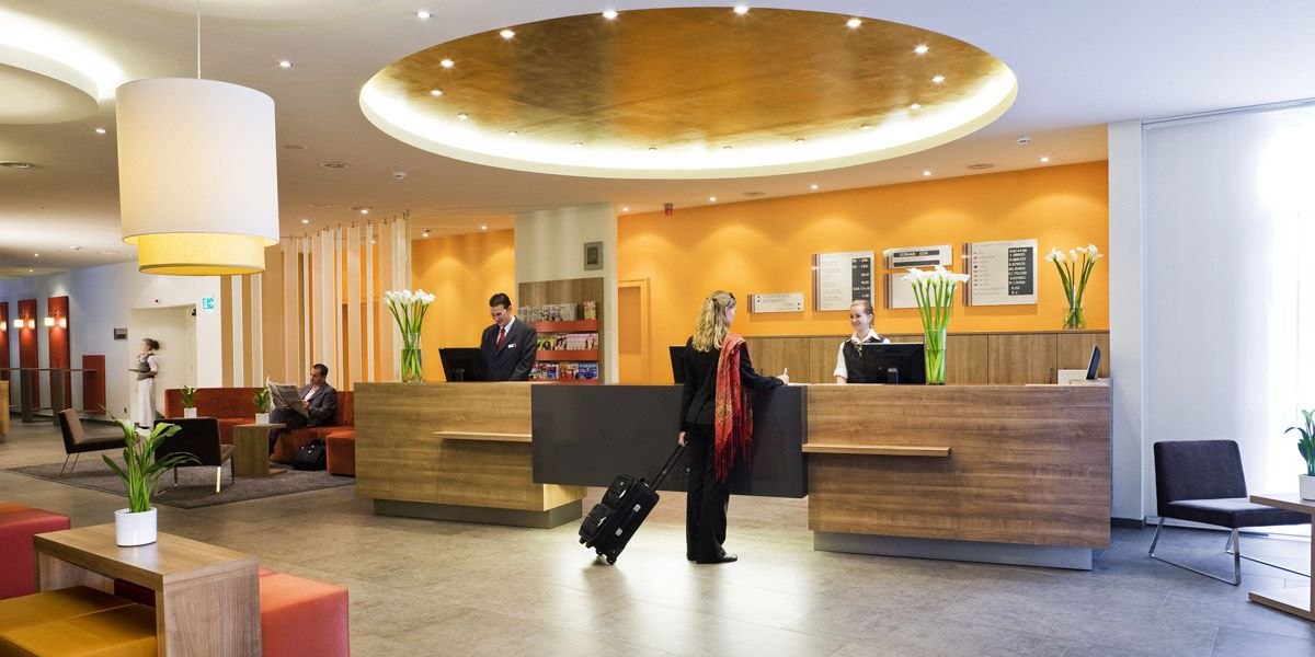 Eine moderne Hotelrezeption mit zwei Angestellten und einer Frau mit Koffer. Im Hintergrund sitzen Gäste auf Sofas. Die Einrichtung ist stilvoll und einladend., © Mercure Stuttgart Airport Messe Eine moderne Hotelrezeption mit zwei Angestellten und einer Frau mit Koffer. Im Hintergrund sitzen Gäste auf Sofas. Die Einrichtung ist stilvoll und einladend., © Mercure Stuttgart Airport Messe