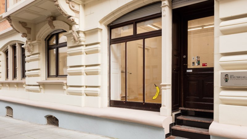 Eingang einer Galerie mit dem Schild 'Die Raumgalerie'. Eine Banane ist an der Glast&uuml;r angebracht. Das Geb&auml;ude hat eine verzierte Fassade., &copy; Dirk Wilhelmy Fotografie