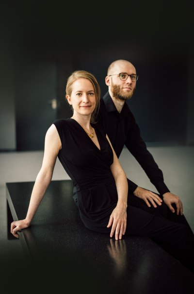 Zwei Personen in eleganter schwarzer Kleidung sitzen auf einem Tisch vor einem dunklen Hintergrund. Sie wirken entspannt und selbstbewusst., © ONYX Klavierduo Zwei Personen in eleganter schwarzer Kleidung sitzen auf einem Tisch vor einem dunklen Hintergrund. Sie wirken entspannt und selbstbewusst., © ONYX Klavierduo