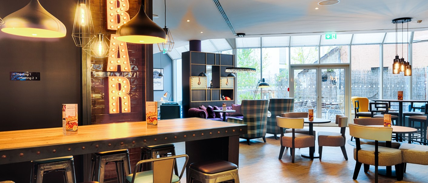 Moderne Bar mit stilvoller Beleuchtung, hohen Tischen und Stühlen. Im Hintergrund gemütliche Sitzbereiche und große Fenster., © Premier Inn Germany