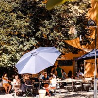 Menschen genie&szlig;en das sonnige Wetter in einem Caf&eacute; im Freien, umgeben von B&auml;umen und Sonnenschirmen. Eine entspannte Atmosph&auml;re herrscht vor., &copy; Stuttgart-Marketing GmbH, Sarah Schmid