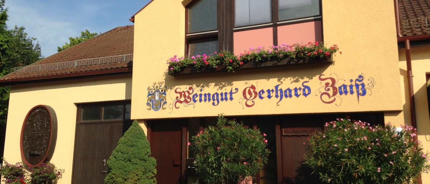 Gelbes Gebäude mit der Aufschrift 'Weingut Gerhard Zaiß', umgeben von Blumen und Pflanzen., © Simone Mack Gelbes Gebäude mit der Aufschrift 'Weingut Gerhard Zaiß', umgeben von Blumen und Pflanzen., © Simone Mack