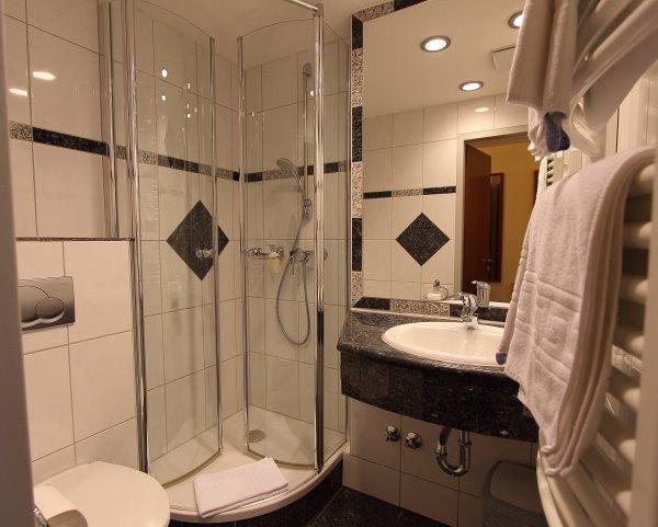 Modernes Badezimmer mit Glasdusche, Waschbecken und Handtuchhalter. Weiße Fliesen mit schwarzen Akzenten. Beleuchtung an der Decke., © Aparthotel Astro