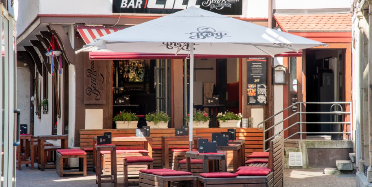 Außenbereich der Café Bar Zill mit Holztischen und roten Sitzpolstern. Ein großer Sonnenschirm spendet Schatten. Im Hintergrund ist der Eingang sichtbar., © Café Bar Zill