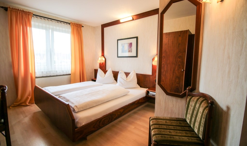 Gemütliches Doppelzimmer mit Holzmöbeln, großem Bett, Spiegel und Fenster mit Vorhängen. Warme Farben und einladende Atmosphäre., © Ulli Kellenbenz Gemütliches Doppelzimmer mit Holzmöbeln, großem Bett, Spiegel und Fenster mit Vorhängen. Warme Farben und einladende Atmosphäre., © Ulli Kellenbenz