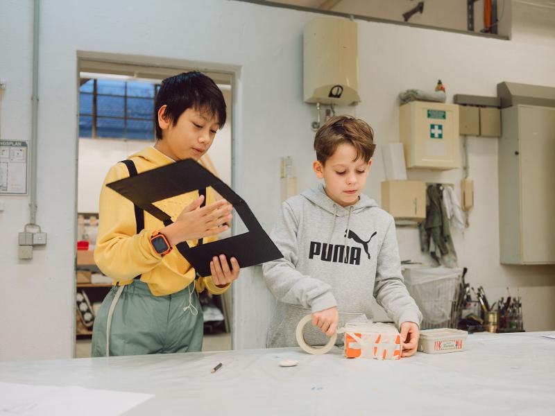Zwei Kinder arbeiten an einem Tisch mit Bastelmaterialien. Eines hält ein schwarzes Papier, das andere benutzt Klebeband. Der Raum wirkt wie eine Werkstatt., © Kunstschule Labyrinth