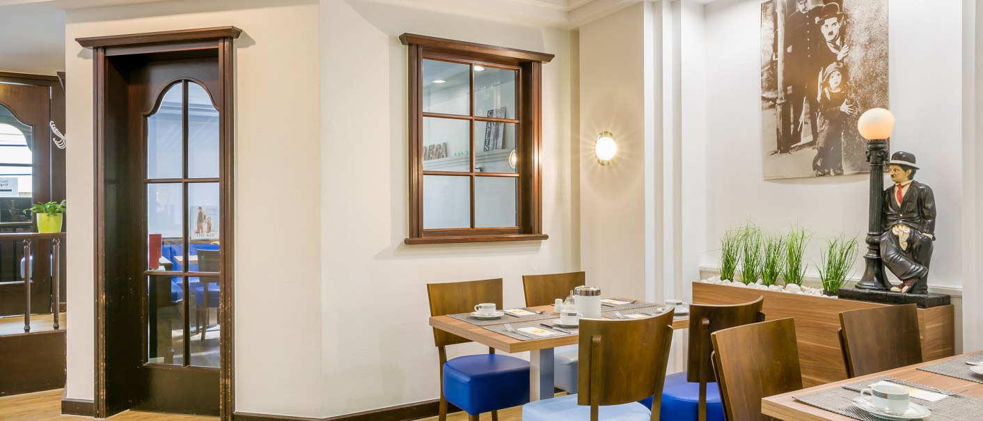 Ein Frühstücksraum mit Holztischen, blauen Stühlen und einer Statue. An der Wand hängt ein Bild, Pflanzen dekorieren den Raum., © TOMAS