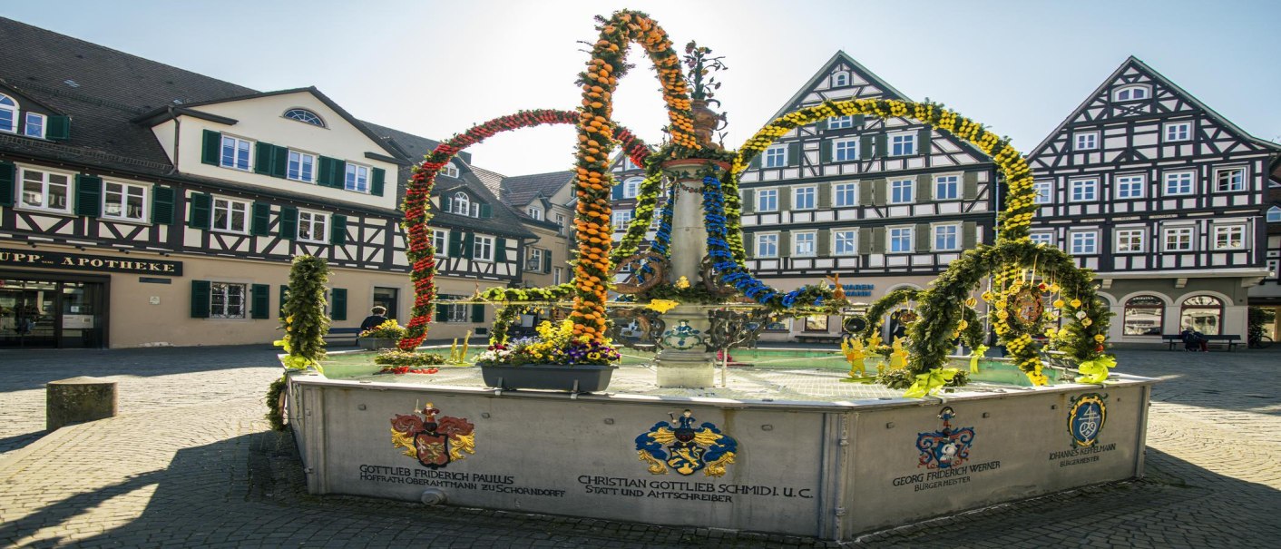 Osterbrunnen auf dem Schorndorfer Marktplatz, geschmückt mit bunten Blumen und Girlanden, umgeben von Fachwerkhäusern., © SMG, Sarah Schmid Osterbrunnen auf dem Schorndorfer Marktplatz, geschmückt mit bunten Blumen und Girlanden, umgeben von Fachwerkhäusern., © SMG, Sarah Schmid
