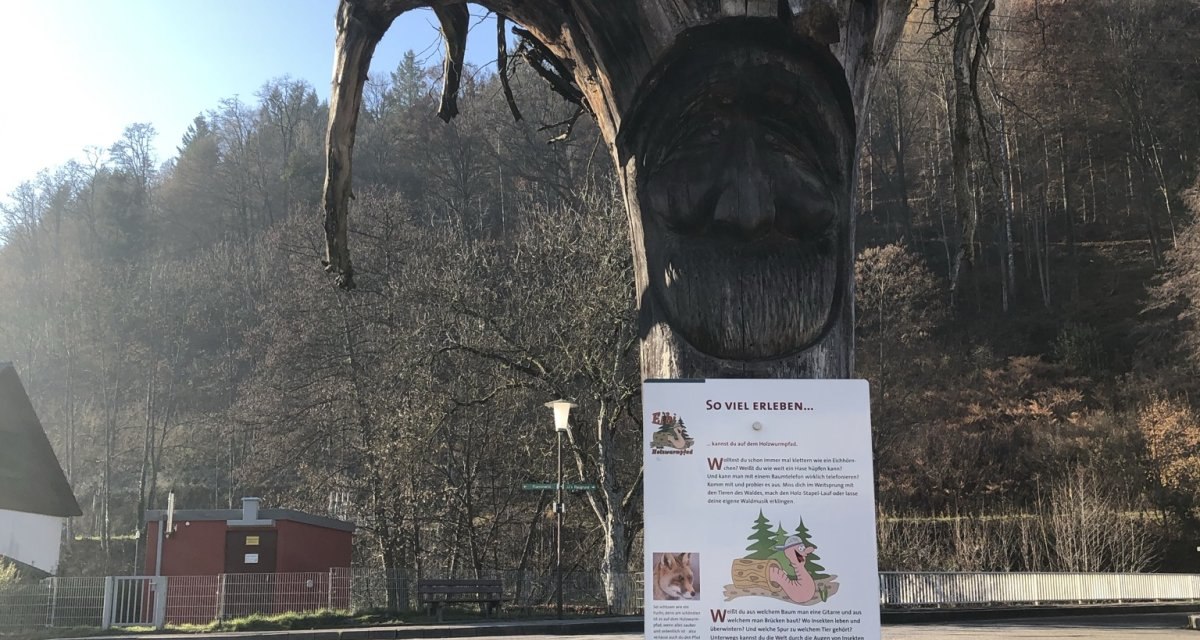 Ein geschnitzter Baumstamm mit Gesicht und Infotafel steht vor einer Stra&szlig;e, umgeben von bewaldeten H&uuml;geln., &copy; N&ouml;rdlicher Schwarzwald