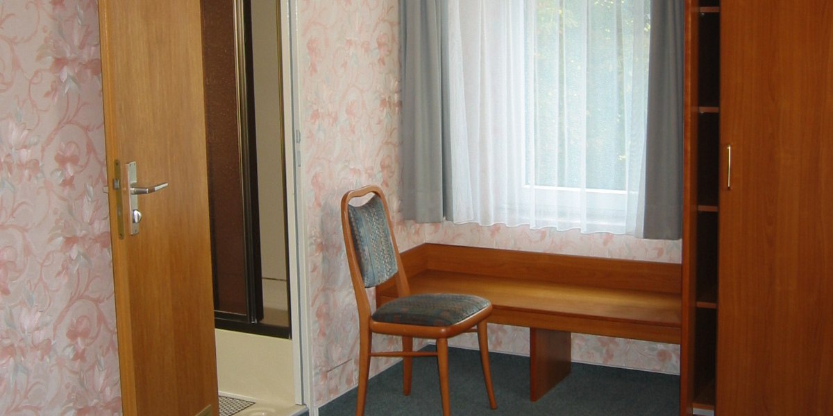 Ein einfach eingerichtetes Hotelzimmer mit einem Stuhl, einem Fenster mit Vorhängen und einer Holzgarderobe., © Hotel Wirt am Berg