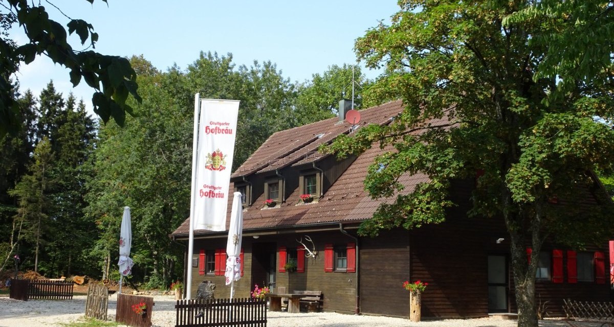 Dunkelbraune Holzhütte mit roten Klappläden im Wald. Vor der Hütte steht eine Fahne und zwei geschlossene Sonnenschirme., © Foto: Frieder Kopper Dunkelbraune Holzhütte mit roten Klappläden im Wald. Vor der Hütte steht eine Fahne und zwei geschlossene Sonnenschirme., © Foto: Frieder Kopper